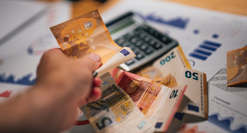 A person’s hand is visible holding a crumpled 10 euro banknote above a cluttered surface with various Euro banknotes, including 20 and 50 euro notes, spread out and partially overlapping. In the background, there is a calculator and printed documents with charts and graphs, likely related to financial or relocation planning. The scene appears to be set inside a property, with the focus on the cash exchange involved in home relocation or moving services. The image emphasizes the financial aspects of house removals, supporting the context of packing, moving logistics, and possible associated costs managed by a company such as Man With a Van Hammersmith, which specializes in removals and transport services.