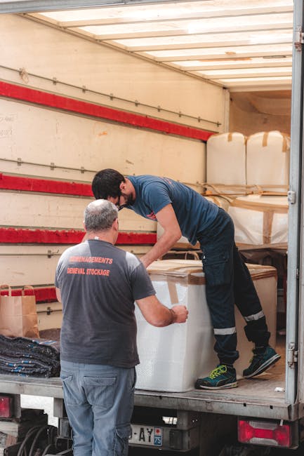 Inside the back of a moving van, two men are engaged in a furniture transport and home relocation process. One man, wearing a dark T-shirt with red and white lettering on the back, is standing on the ground, holding a large white cardboard box wrapped with brown packing tape. The second man, dressed in a dark blue T-shirt and black trousers with reflective strips, is standing on the van's loading area, leaning slightly forward with one knee bent, as he guides the box onto the vehicle. The van's interior features a wooden floor, a metal rail with red stripes along the side wall, and a partially visible ceiling. In the background, there are several large white packages wrapped in plastic or fabric and secured with brown tape, stacked against the van’s wall. Some loose packing materials and a black plastic bag are visible nearby, indicating a packing and moving operation typical of professional removals services offered by companies like Man With a Van Hammersmith. Proper lighting illuminates the scene, capturing the detailed actions involved in loading furniture and boxes for a move.