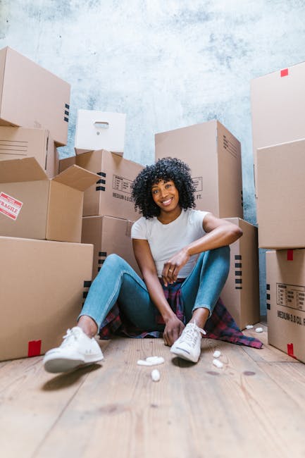 A young woman with curly black hair, wearing a white t-shirt and light blue jeans, is sitting on a wooden floor surrounded by numerous cardboard boxes of various sizes, some sealed with packing tape and others open, revealing packing materials. She is smiling and relaxed while interior natural light illuminates the space, which has a light blue wall with a textured finish. The boxes are stacked and arranged around her, indicating a home relocation or packing process. Nearby, a few crumpled packing papers are on the floor. This scene reflects furniture transport and packing activities involved in professional house removals. The setting suggests an organized move, possibly managed by Man With a Van Hammersmith, a company specialising in removals in the Queen Caroline Estate W6 area.
