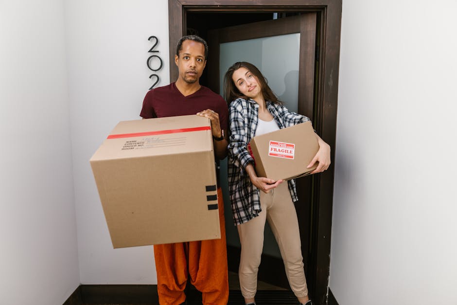 A young woman with curly black hair, wearing a white t-shirt and light blue jeans, is sitting on a wooden floor surrounded by numerous cardboard boxes of various sizes, some sealed with packing tape and others open, revealing packing materials. She is smiling and relaxed while interior natural light illuminates the space, which has a light blue wall with a textured finish. The boxes are stacked and arranged around her, indicating a home relocation or packing process. Nearby, a few crumpled packing papers are on the floor. This scene reflects furniture transport and packing activities involved in professional house removals. The setting suggests an organized move, possibly managed by Man With a Van Hammersmith, a company specialising in removals in the Queen Caroline Estate W6 area.