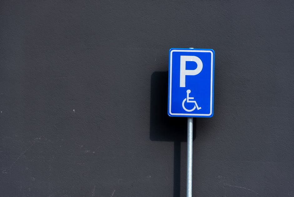 A blue wheelchair-accessible parking sign mounted on a metal post is positioned against a dark grey exterior wall. The sign displays a white letter 'P' and a symbol of a person in a wheelchair, indicating designated parking for disabled individuals. In the background, the pavement is visible at the base of the wall, with shadows cast by the sign suggesting a sunny day. This scene relates to parking regulations and permits for house removals, possibly indicating the location where a vehicle from Man With a Van Hammersmith is preparing for furniture transport during a home relocation process, aligned with the house removals and moving services discussed on the page about Hammersmith and Fulham council rules.