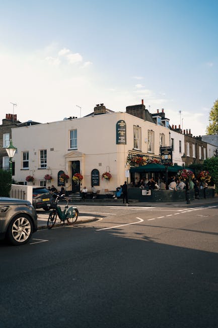 An exterior view of a two-storey building on a city street during daytime, with a white facade and multiple windows adorned with flower boxes. The ground floor features a cafe or restaurant with outdoor seating, a dark green awning, and people sitting at tables. In front of the building, a black van is parked on the road, and a bicycle is secured to a bike rack near the sidewalk. A person is walking on the pavement, and a few others are visible seated outside the establishment. The street has marked lanes and a pedestrian crossing. The sky above is mostly clear with some clouds, and there is a lamppost on the corner. This setting reflects typical urban surroundings suitable for residential or commercial properties involved in home relocation or furniture transport, supported by house removals specialists like Man With a Van Hammersmith.