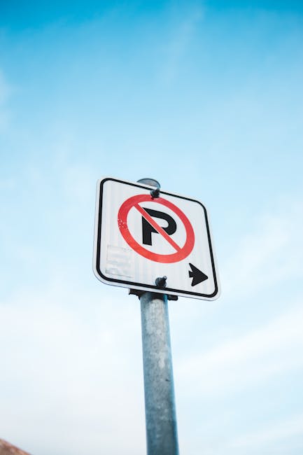 A blue wheelchair-accessible parking sign mounted on a metal post is positioned against a dark grey exterior wall. The sign displays a white letter 'P' and a symbol of a person in a wheelchair, indicating designated parking for disabled individuals. In the background, the pavement is visible at the base of the wall, with shadows cast by the sign suggesting a sunny day. This scene relates to parking regulations and permits for house removals, possibly indicating the location where a vehicle from Man With a Van Hammersmith is preparing for furniture transport during a home relocation process, aligned with the house removals and moving services discussed on the page about Hammersmith and Fulham council rules.