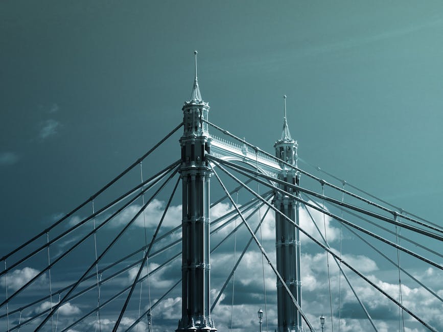 A detailed view of the towers and suspension cables of Hammersmith Bridge, with two tall, ornate pinnacles visible at the top of each tower. The bridge features numerous steel cables extending from the towers, creating a web-like pattern against a partly cloudy sky. The image captures the structural elements and architectural details of the historic suspension bridge, emphasizing its precision engineering and decorative features. The scene is taken during daytime, with natural light highlighting the metallic surfaces and the sky providing a contrasting background. This prominent bridge is associated with areas such as Hammersmith and may feature in residential or commercial property relocation services, where careful transportation of furniture and equipment through or around the bridge may be involved. Man With a Van Hammersmith utilises careful planning and appropriate equipment to facilitate home relocation or furniture transport over such structures, supporting efficient moving and packing operations in the London area.