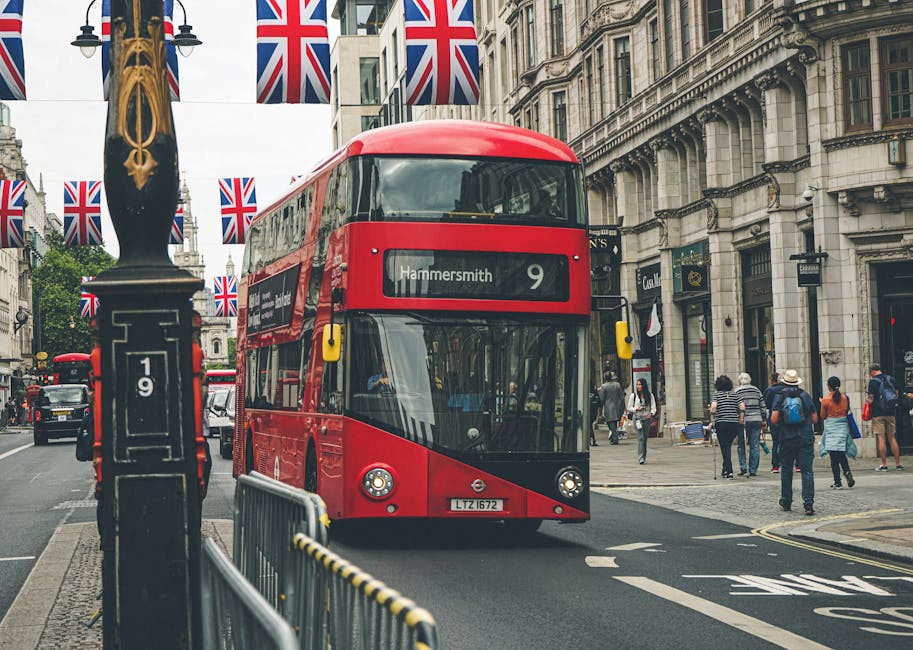 A red double-decker bus with destination Hammersmith is traveling down a busy city street with pedestrians walking on the sidewalk on both sides. The street is lined with historic and modern buildings, some of which have decorative stone facades and large windows. British flags are hanging from lampposts along the street, enhancing the festive or patriotic atmosphere. In the foreground, a metal barrier is placed on the pavement, and a black lamppost with gold decorative elements stands nearby. Within the context of home relocation or furniture transport, this image depicts the typical urban environment where a professional removals company like Man With a Van Hammersmith might operate, facilitating the logistics involved in packing, moving, and delivering household items across the W6 postcode area. The scene captures the movement and flow of city life, illustrating the challenges of navigating busy streets during a furniture transport or home moving service.