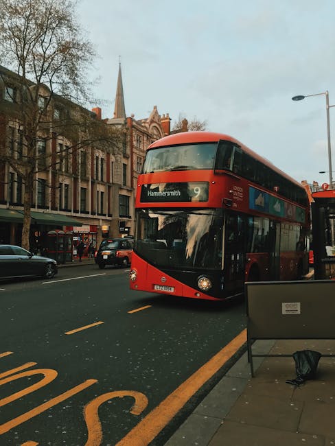 A red double-decker bus traveling along a street in Hammersmith, London, with a destination display indicating route 9 towards Hammersmith. The bus is approaching a bus stop with a bench and a black bag placed on the ground. Behind the bus, there are multi-storey buildings with large windows and a church or chapel with a tall, pointed steeple visible against a cloudy sky. Several parked cars and a few pedestrians are seen on the sidewalk, which is lined with leafless trees, suggesting late autumn or winter. The scene captures an urban environment typical for house removals and furniture transport, demonstrating the busy street scene where professionals like Man With a Van Hammersmith conduct home relocation or moving logistics amidst city traffic.