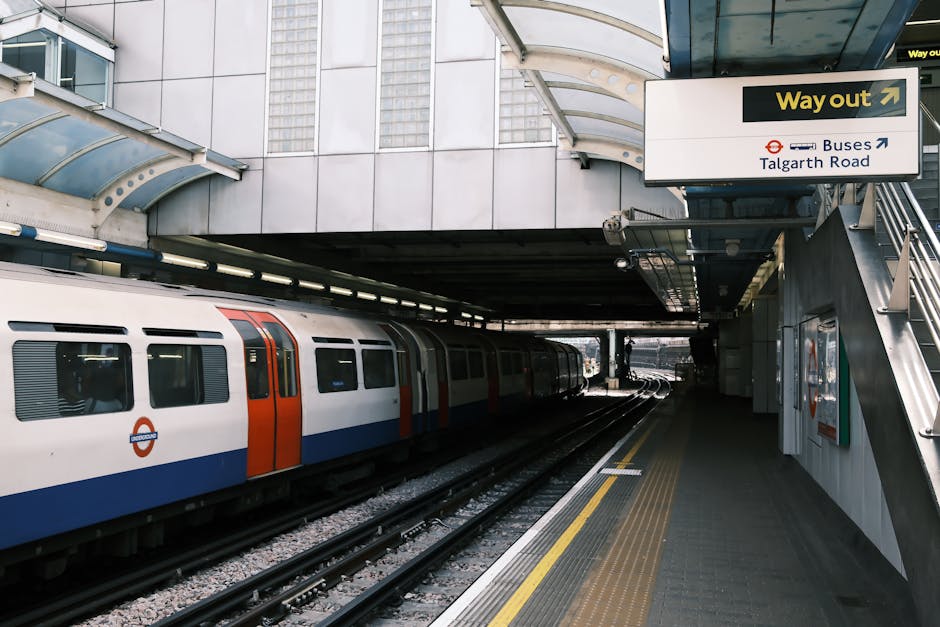 Inside a modern London underground station, a white and orange train is stationary alongside platform level, with multiple windows and doors visible. The platform is equipped with a tactile paving strip near the edge for safety, and the surface is paved with dark tiles. Overhead, a large rectangular sign with a black arrow and the words 'Way out' indicates the exit to Taltagh Road and bus connections, mounted on a grey metallic structure. To the right, there is a staircase with metal railings leading upward, and behind it, a poster or informational display is partially visible on the wall. The station environment is lit with natural daylight filtering through the station roof, which features a curved, translucent canopy made from glass or plastic panels supported by metal framework. This setting depicts the typical scene of a public transport embarkation or disembarkation point, relevant to home relocation or moving logistics involving transport connections to suburban or urban areas, with visible focus on travel infrastructure.