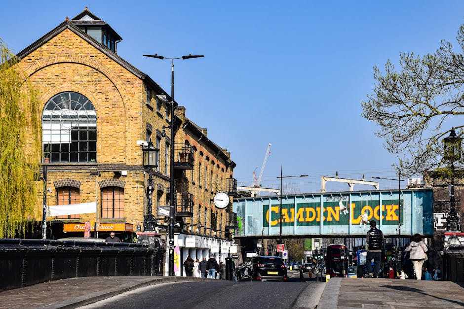 A red double-decker bus traveling along a street in Hammersmith, London, with a destination display indicating route 9 towards Hammersmith. The bus is approaching a bus stop with a bench and a black bag placed on the ground. Behind the bus, there are multi-storey buildings with large windows and a church or chapel with a tall, pointed steeple visible against a cloudy sky. Several parked cars and a few pedestrians are seen on the sidewalk, which is lined with leafless trees, suggesting late autumn or winter. The scene captures an urban environment typical for house removals and furniture transport, demonstrating the busy street scene where professionals like Man With a Van Hammersmith conduct home relocation or moving logistics amidst city traffic.