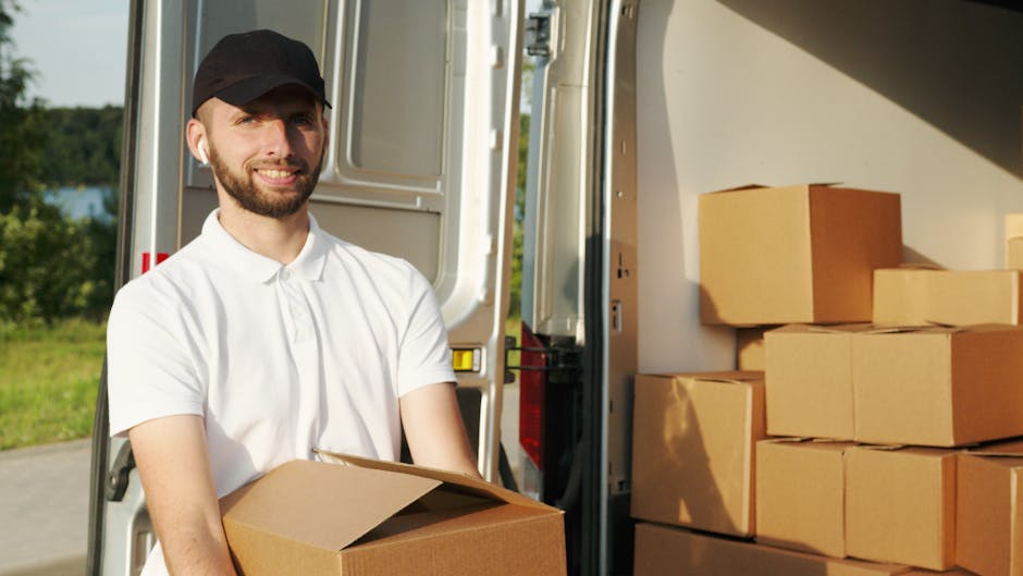 A male mover from Man With a Van Hammersmith, wearing a white polo shirt and a black cap, is standing outside a van during the loading process. He is holding a medium-sized cardboard box and smiling at the camera. The van, parked on a paved area, has its rear door open, revealing multiple brown cardboard boxes neatly stacked inside and on the pavement nearby. The background shows green trees and a clear sky, indicating an outdoor setting. The scene captures a home relocation or furniture transport operation, with packing materials and boxes ready for moving. The vehicle appears to be a small to medium-sized moving van, equipped to facilitate efficient packing and moving services, aligned with the services offered by [COMPANY_NAME] in house removals and packing logistics.