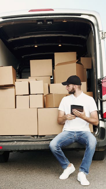 A man dressed in a white t-shirt, blue jeans, and a black cap is sitting on the edge of an open van in Hammersmith, with the vehicle loaded with numerous cardboard boxes of varying sizes, some sealed and others open, revealing packing materials like plastic wrap and paper. The boxes are stacked inside the van, which is parked on a paved surface, possibly at a property or loading area. The man is holding a tablet device, likely coordinating or checking a home relocation or furniture transport process. The interior of the van shows a dark roof with some lighting fixtures, and the environment appears bright, suggesting daytime. The scene reflects a moving or packing operation related to house removals, with the focus on loading, packing, and preparing furniture and belongings for transportation, aligning with professional removal services offered by Man With a Van Hammersmith.