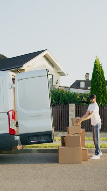 A male mover from Man With a Van Hammersmith, wearing a white polo shirt and a black cap, is standing outside a van during the loading process. He is holding a medium-sized cardboard box and smiling at the camera. The van, parked on a paved area, has its rear door open, revealing multiple brown cardboard boxes neatly stacked inside and on the pavement nearby. The background shows green trees and a clear sky, indicating an outdoor setting. The scene captures a home relocation or furniture transport operation, with packing materials and boxes ready for moving. The vehicle appears to be a small to medium-sized moving van, equipped to facilitate efficient packing and moving services, aligned with the services offered by [COMPANY_NAME] in house removals and packing logistics.