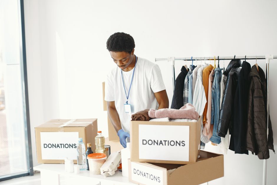 A young man wearing a white t-shirt, blue gloves, and a lanyard is inside a room with white walls, engaging in a home relocation or packing process. He is standing behind a table that holds three card