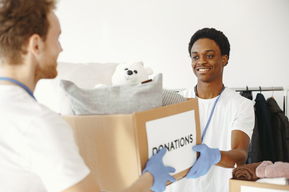 A young man wearing a white t-shirt, blue gloves, and a lanyard is inside a room with white walls, engaging in a home relocation or packing process. He is standing behind a table that holds three card
